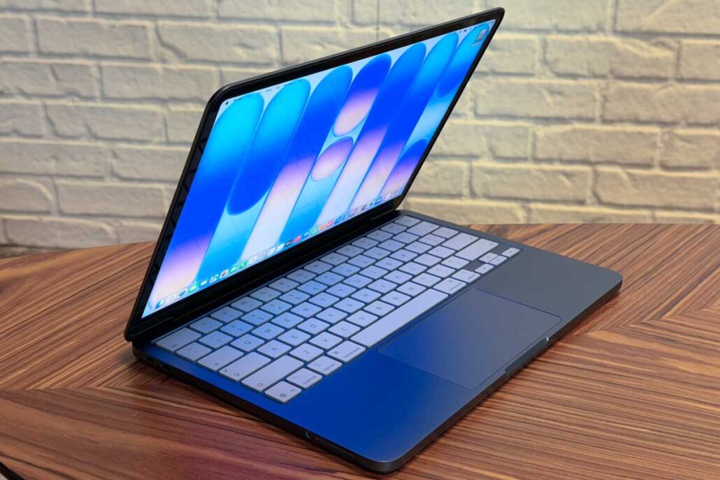 Open laptop on a wooden table, displaying a blue abstract wallpaper with a white keyboard and trackpad, brick wall in the background.