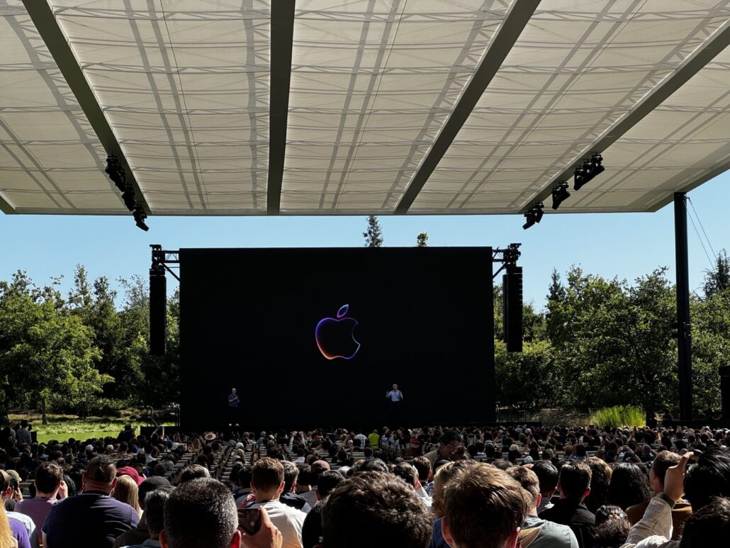 Outdoor tech presentation with a large black stage screen displaying the Apple logo and a crowd watching.