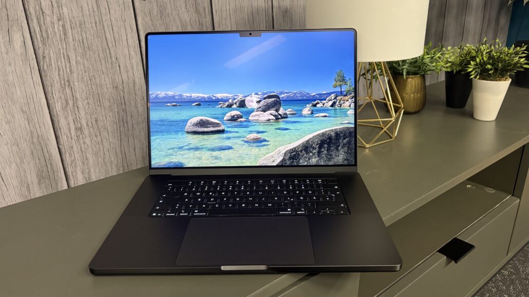 Laptop on a gray desk showing a tropical seascape wallpaper with rocks and turquoise water; background includes a lamp and potted plants.