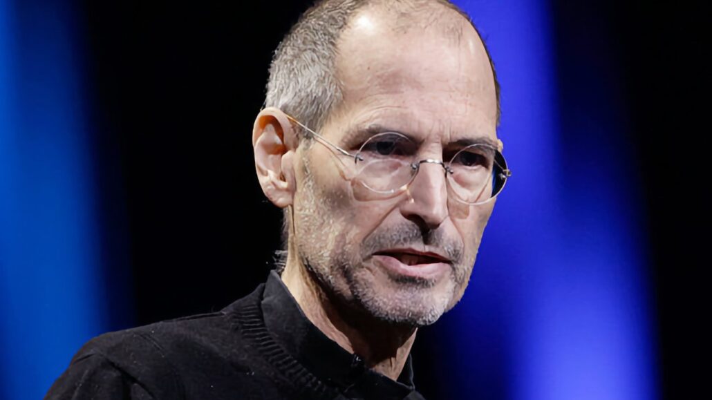 Steve Jobs made his last WWDC appearance 15 years ago today - Dans Tutorials - Dans Tutorials Portrait of a middle-aged man with glasses and stubble, wearing a black shirt, speaking on a stage with blue lighting in the background.