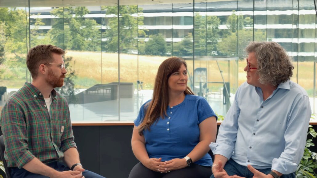 Three coworkers seated in a bright glass-walled office, conversing.