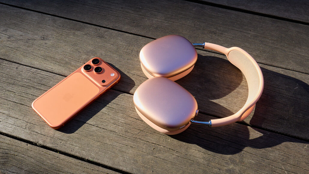Rose gold iPhone with triple cameras beside pink over-ear wireless headphones on a wooden surface.