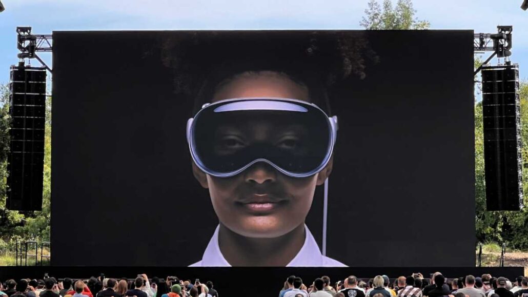 Large outdoor screen shows a person wearing a virtual reality headset; crowd seated in front watching the display.