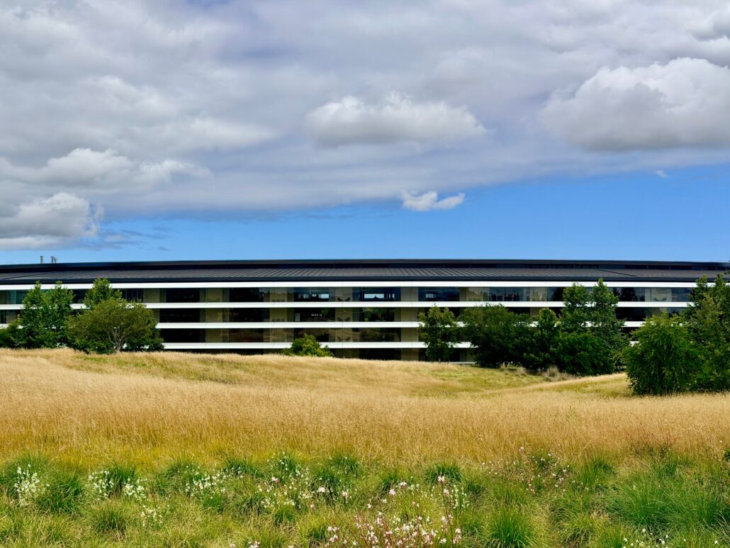 Modern low-rise glass office building set in a grassy, semi-arid field with trees in the foreground under a cloudy sky.